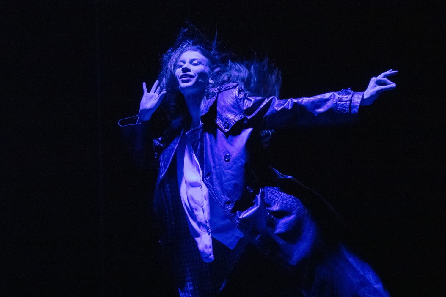 A female dancer with wild hair stands in stark blue light, a hand to her ear as she listens with a serene smile.