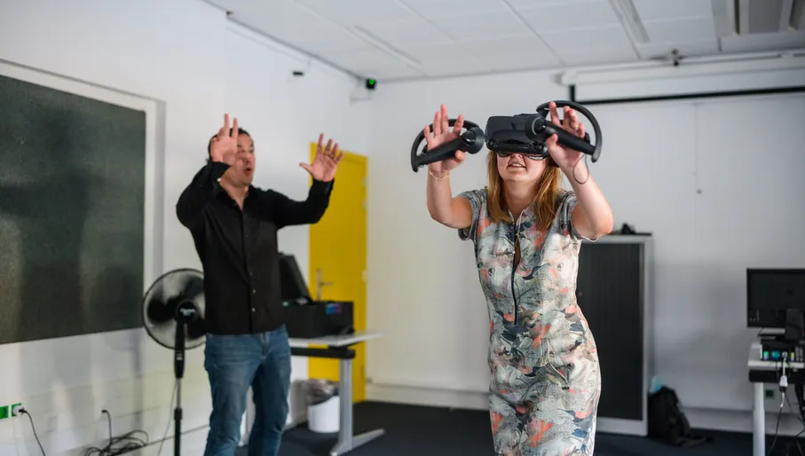 A woman wearing a VR headset and hand-held controllers raises her hands, while a man looks on gesturing at her.
