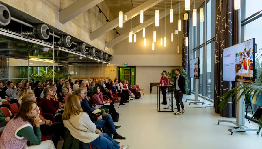 Long shot of a talk in a modern Amsterdam building, with multiple screens hsowing a presentation behind a speaker facing a seated crowd.
