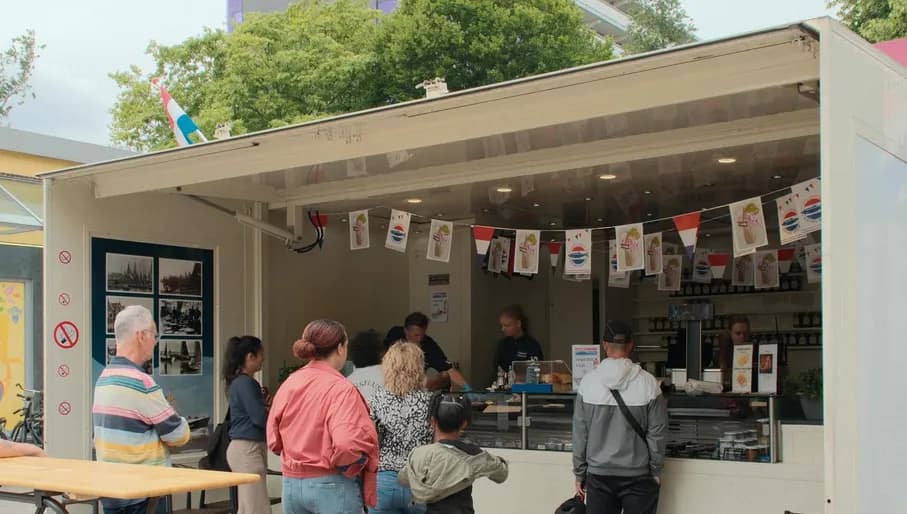 The photo was taken at Molenwijk 23 in Amsterdam-Noord and shows Vishandel De Boer & Klouwer. It shows a classic Dutch herring stall, with a few people waiting to be served by the staff behind the counter. The stall is decorated with small Dutch flags and photos of boats.