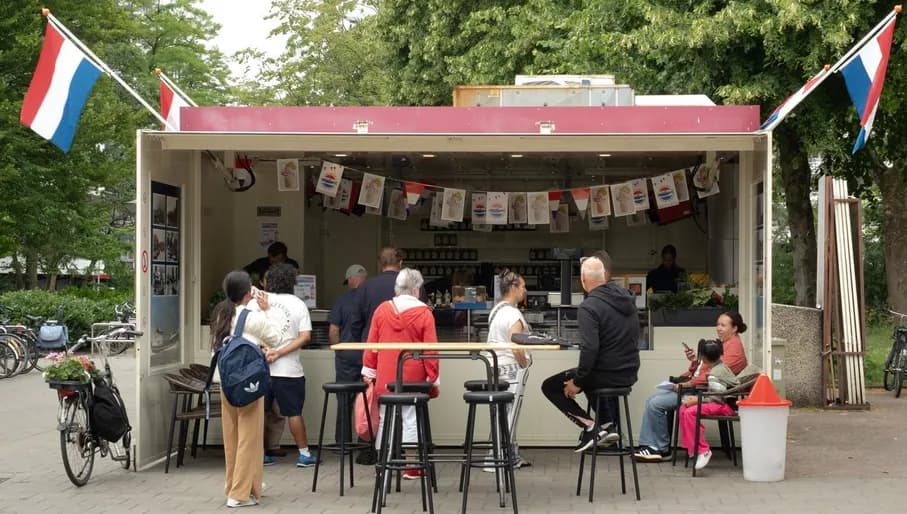The photo was taken at Molenwijk 23 in Amsterdam-Noord and shows Vishandel De Boer & Klouwer. It shows a classic Dutch herring stall, with a few people waiting to be served by the staff behind the counter. The stall is decorated with small Dutch flags and photos of boats and two big Dutch flags on each side. There is also a bike on the left side of the stall.