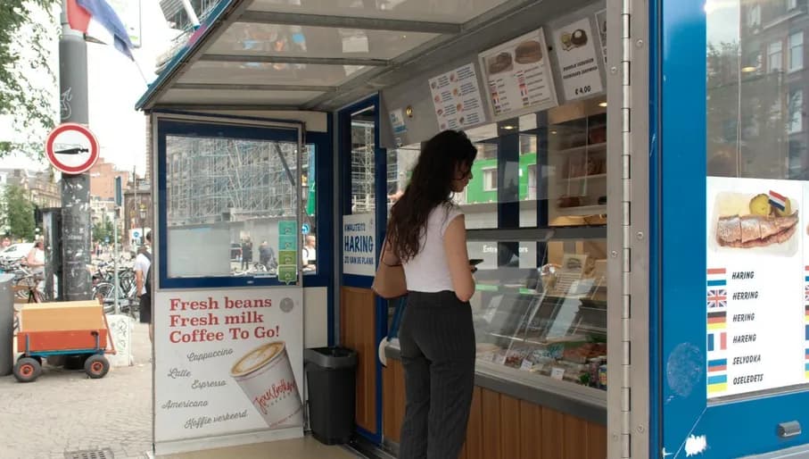 The photo was taken at Nieuwezijds Voorburgwal 200 in Amsterdam-Centrum, behind Dam Square, and shows herring stall Haring & Zo. It captures a woman waiting to be served.
