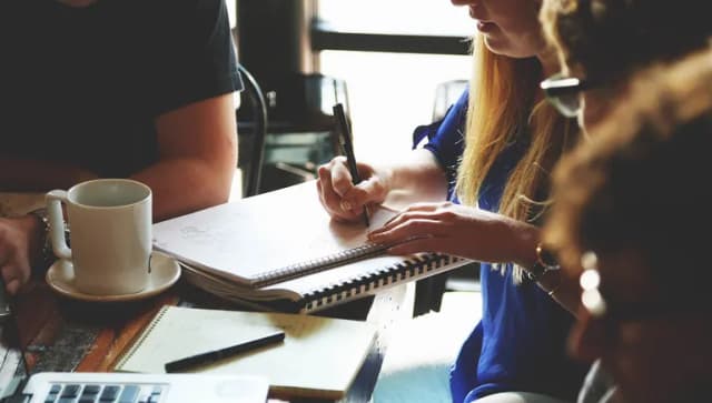 Closeup of hands taking notes on pad of paper next to coffee cup at desk during meeting.