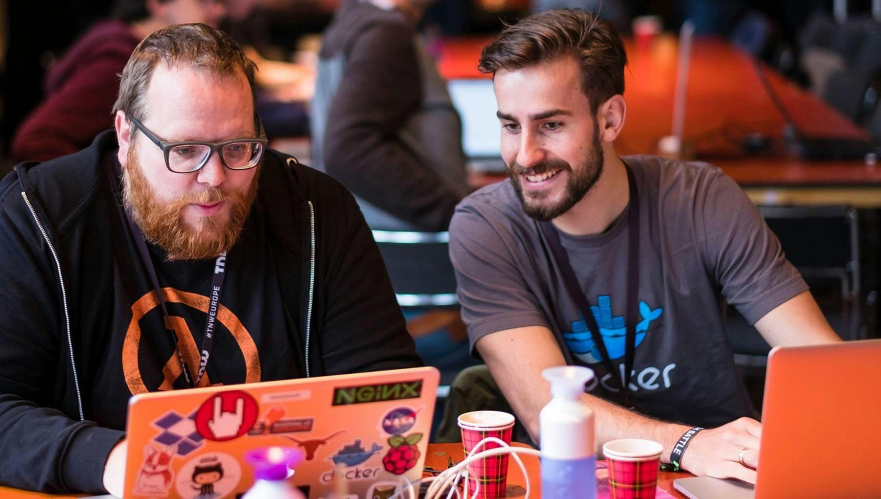 Two people seated with their laptops at The Next Web Amsterdam 2016 Hack Battle, one man smiling as he looks at another's screen.