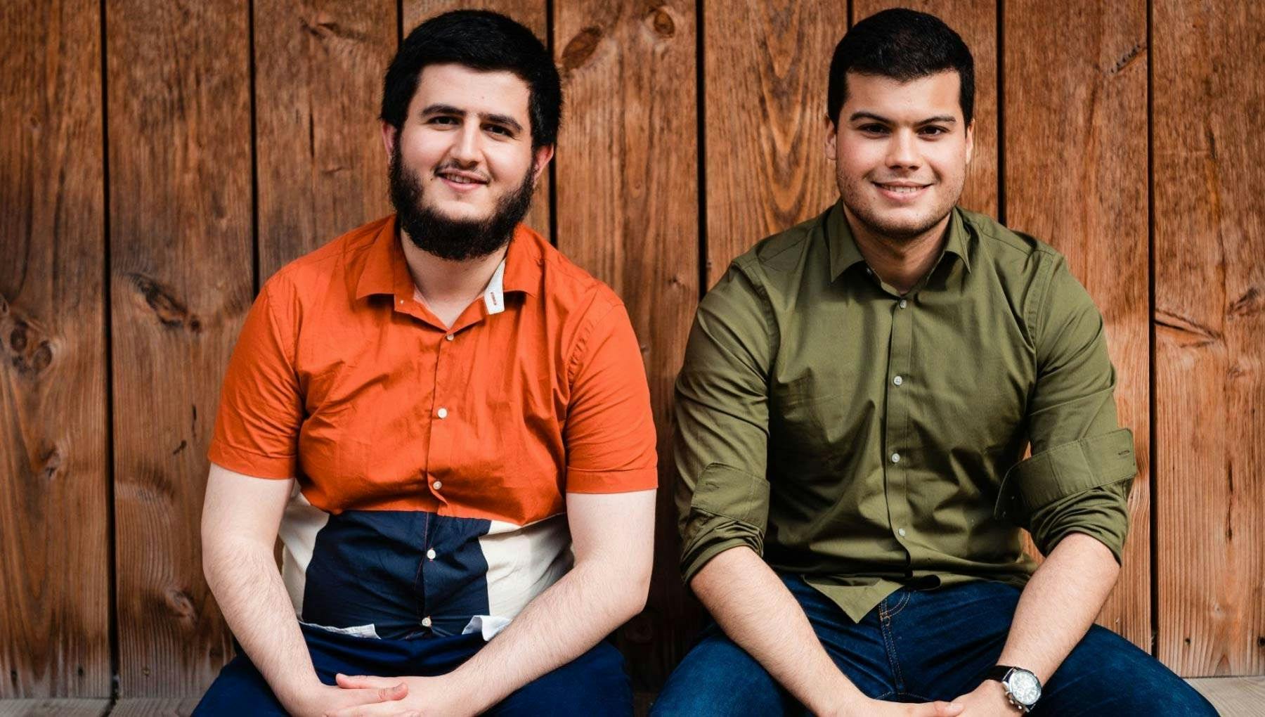 Landscape portrait of Adil Ayi and Marwan el Morabet, co-founders of Omnia, sitting with hands in laps against wooden wall.
