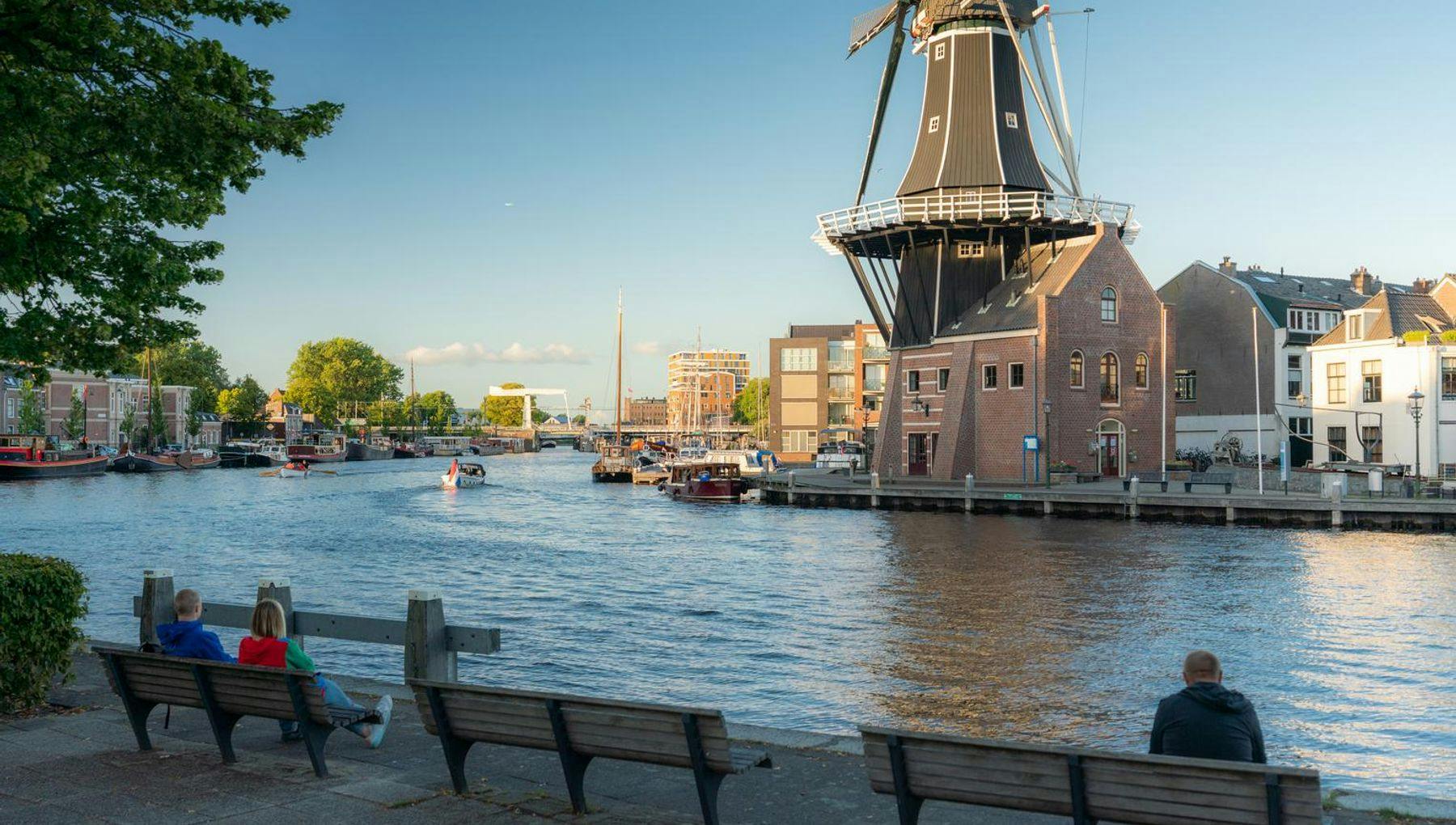 People sitting on benches enjoying their view at the Spaarne river in Haarlem. On the other side of the water, you see the Molen De Adriaan windmill.