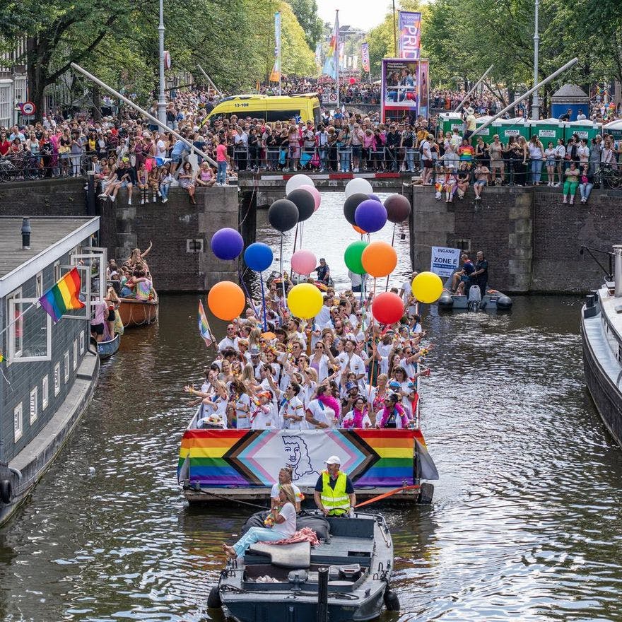 A crowd of people on the streets and in a boat with flags and signs - Pride Canal Parade