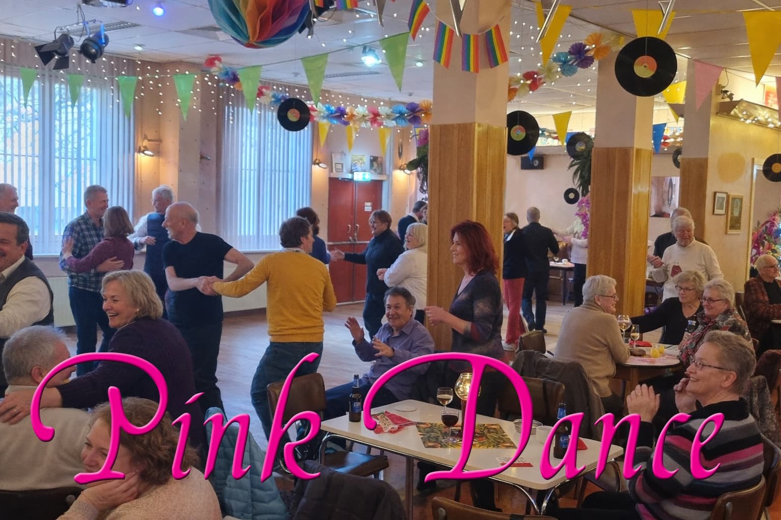 Een levendige dansscène van Pink Dance in Het Open Huis. Een diverse groep dansers – jong en oud, mannen en vrouwen in kleurrijke kleding – beweegt enthousiast op de dansvloer met ballroom- en Latin-stijlen.