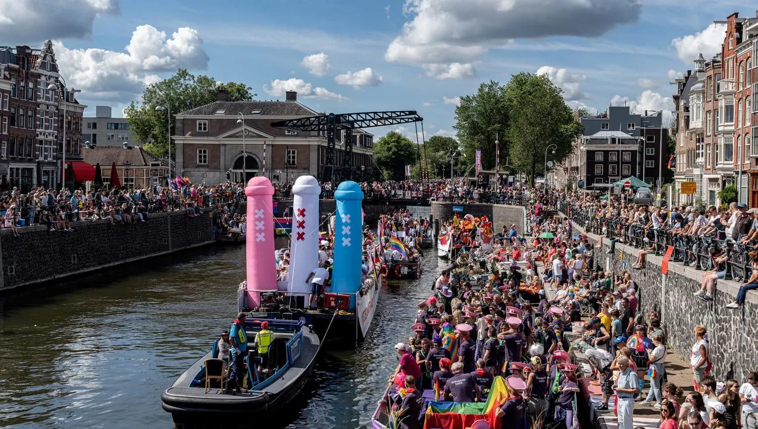 A crowd of people on the streets and in a boat with flags and signs - Pride Canal Parade