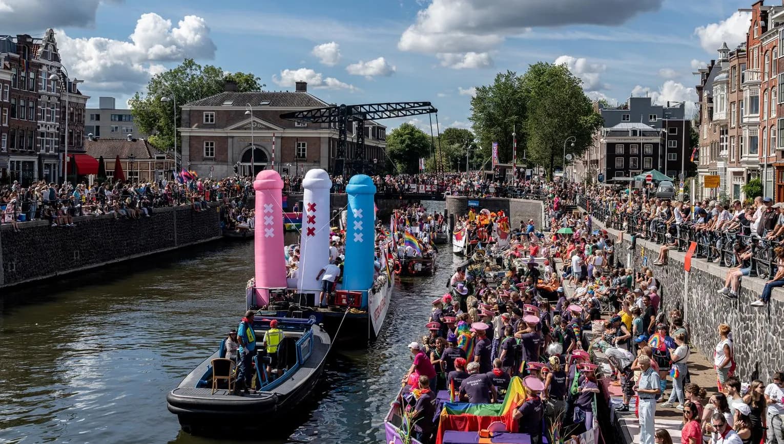 A crowd of people on the streets and in a boat with flags and signs - Pride Canal Parade