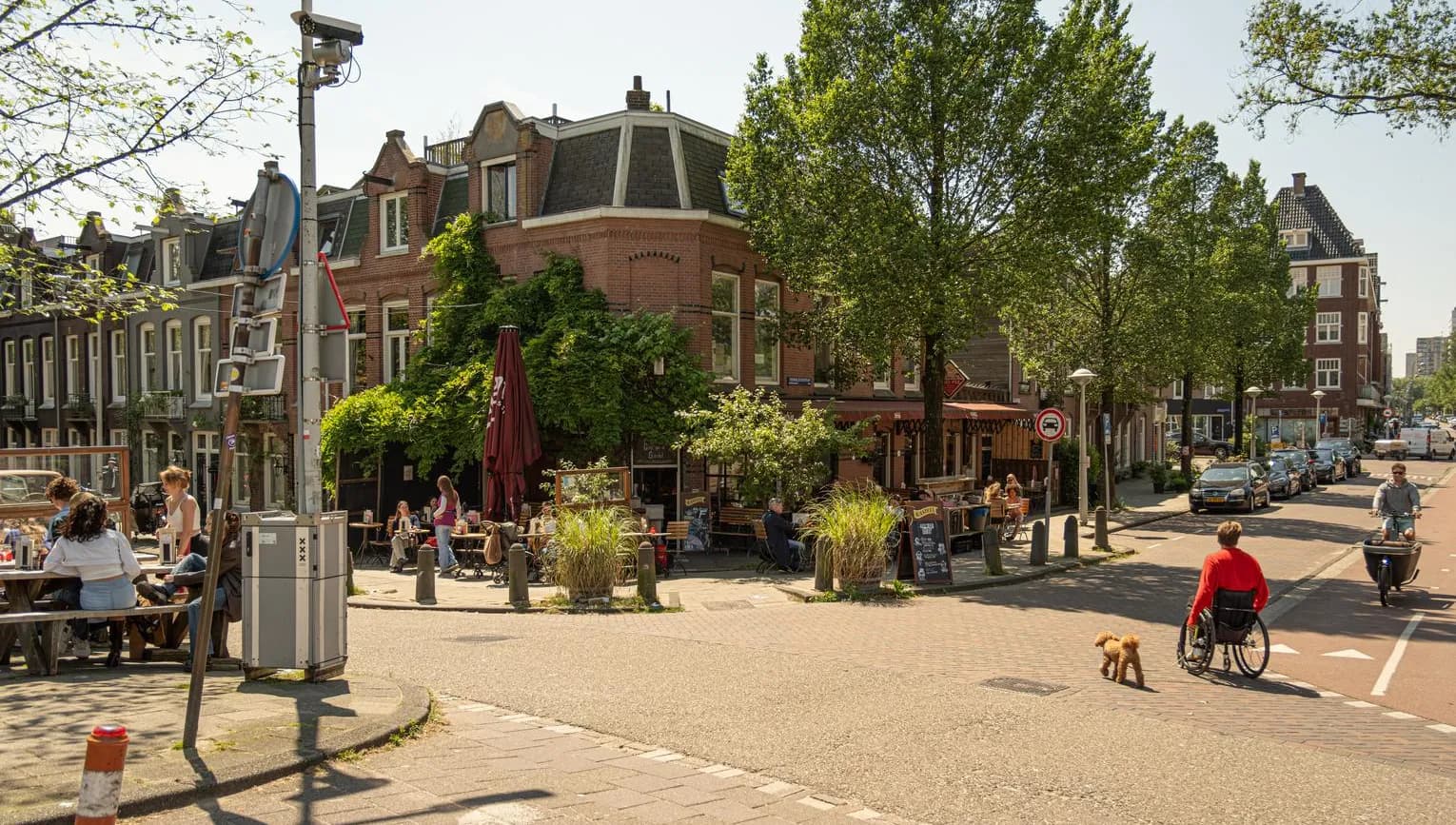 Cyclist and wheelchair user with dog crossing road towards restaurant terrace on Schinkelhavenstraat