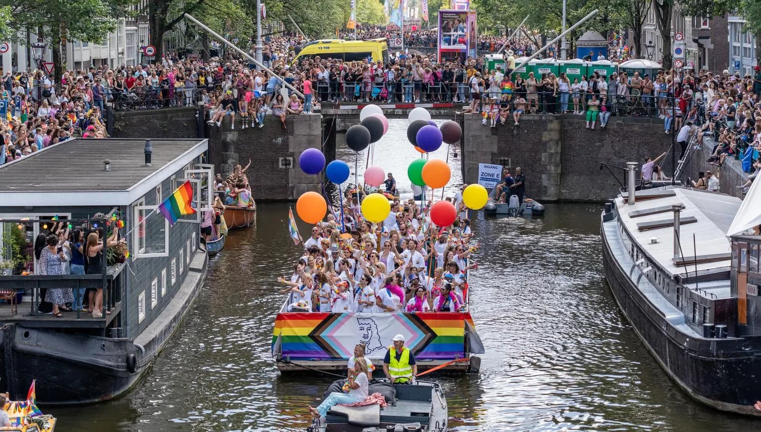 A crowd of people on the streets and in a boat with flags and signs - Pride Canal Parade
