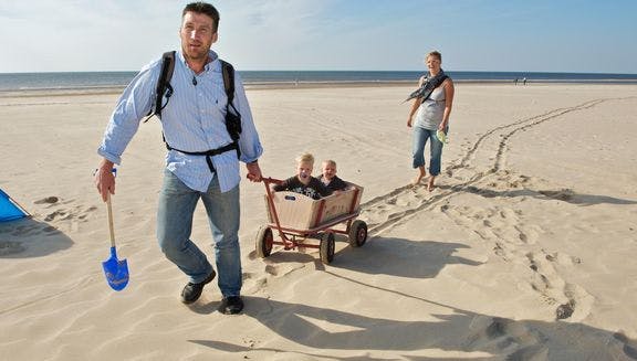 A father is pulling his kids in a handcart over the beach, while the mother follows behind.