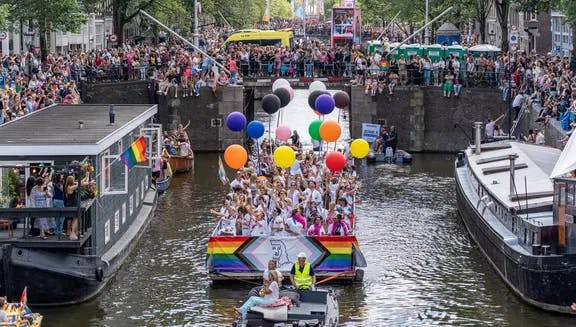 A crowd of people on the streets and in a boat with flags and signs - Pride Canal Parade