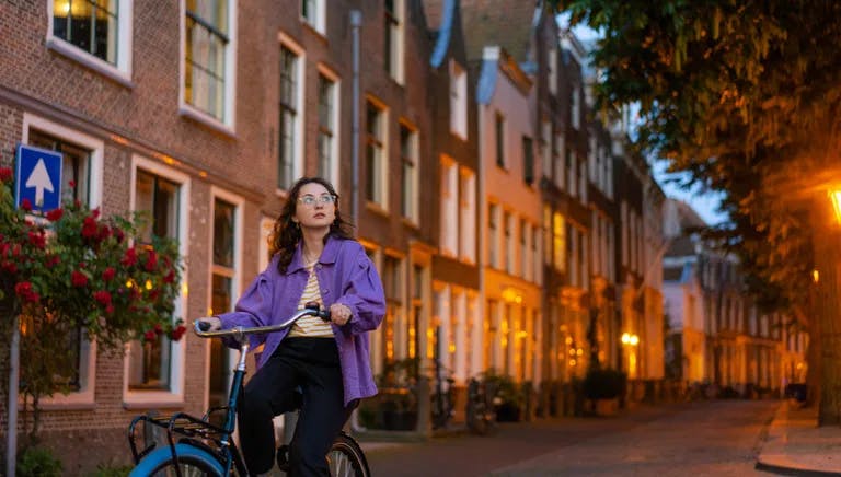 Young Caucasian woman in purple coat riding bicycle in the  city at dusk