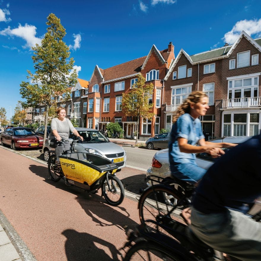 People riding bikes on Haarlem street with woman riding yellow shared Cargoroo e-cargo bike elektrische buurtbakfiets - fast commuting on Dutch street