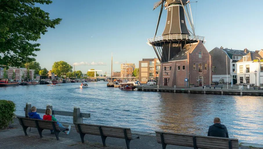People sitting on benches enjoying their view at the Spaarne river in Haarlem. On the other side of the water, you see the Molen De Adriaan windmill.